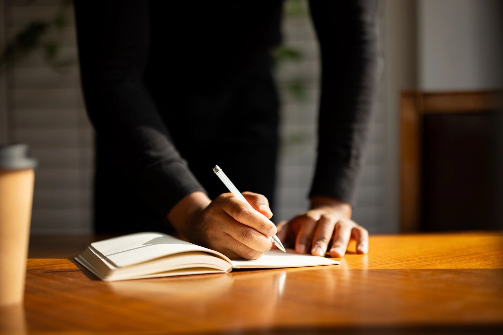 Person writing notes in an open notebook on a wooden desk with a coffee cup nearby